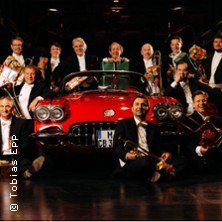 Eine Gruppe von Männern in Anzügen posiert fröhlich vor einem roten Oldtimer, einige halten Musikinstrumente. Der Hintergrund ist dunkel., © links im Bild Eine Gruppe von Männern in Anzügen posiert fröhlich vor einem roten Oldtimer, einige halten Musikinstrumente. Der Hintergrund ist dunkel., © links im Bild