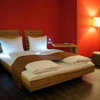 Modernes Hotelzimmer mit roter Wand, Doppelbett, Nachttischlampen und Stehlampe. Elegante Einrichtung mit Holzmöbeln und warmem Licht., © AMH Airport-Messe-Hotel