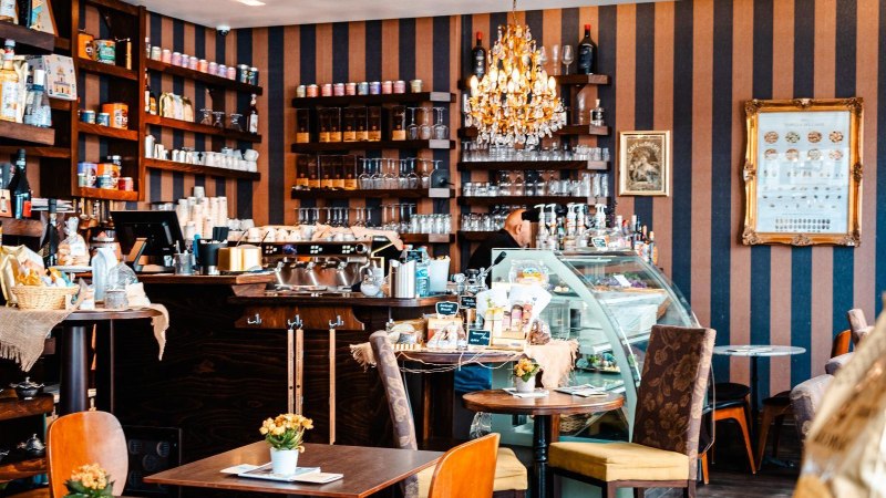 Gemütliches Kaffeehaus mit Kronleuchter, Regalen voller Produkte und einer Theke mit Kaffeemaschine. Tische mit Blumen dekoriert., © Stuttgart-Marketing GmbH, Sarah Schmid Gemütliches Kaffeehaus mit Kronleuchter, Regalen voller Produkte und einer Theke mit Kaffeemaschine. Tische mit Blumen dekoriert., © Stuttgart-Marketing GmbH, Sarah Schmid