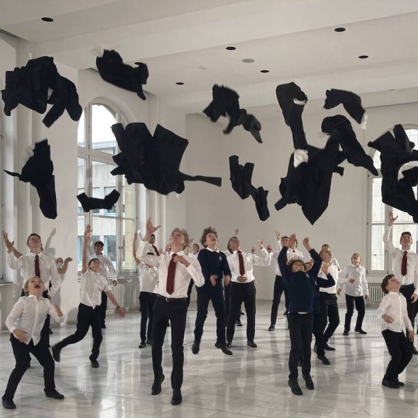 Junge Sänger in weißen Hemden und roten Krawatten werfen schwarze Jacken in die Luft in einem hellen Raum mit großen Fenstern., © Stuttgarter Hymnus-Chorknaben