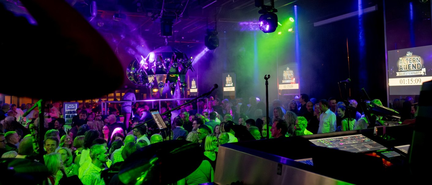 Eine lebhafte Party mit vielen Menschen, bunten Lichtern und einem DJ-Pult im Vordergrund. Im Hintergrund sind Bildschirme mit "Älternabend Silvesterparty" zu sehen., © Adventure Perkins GmbH Eine lebhafte Party mit vielen Menschen, bunten Lichtern und einem DJ-Pult im Vordergrund. Im Hintergrund sind Bildschirme mit "Älternabend Silvesterparty" zu sehen., © Adventure Perkins GmbH