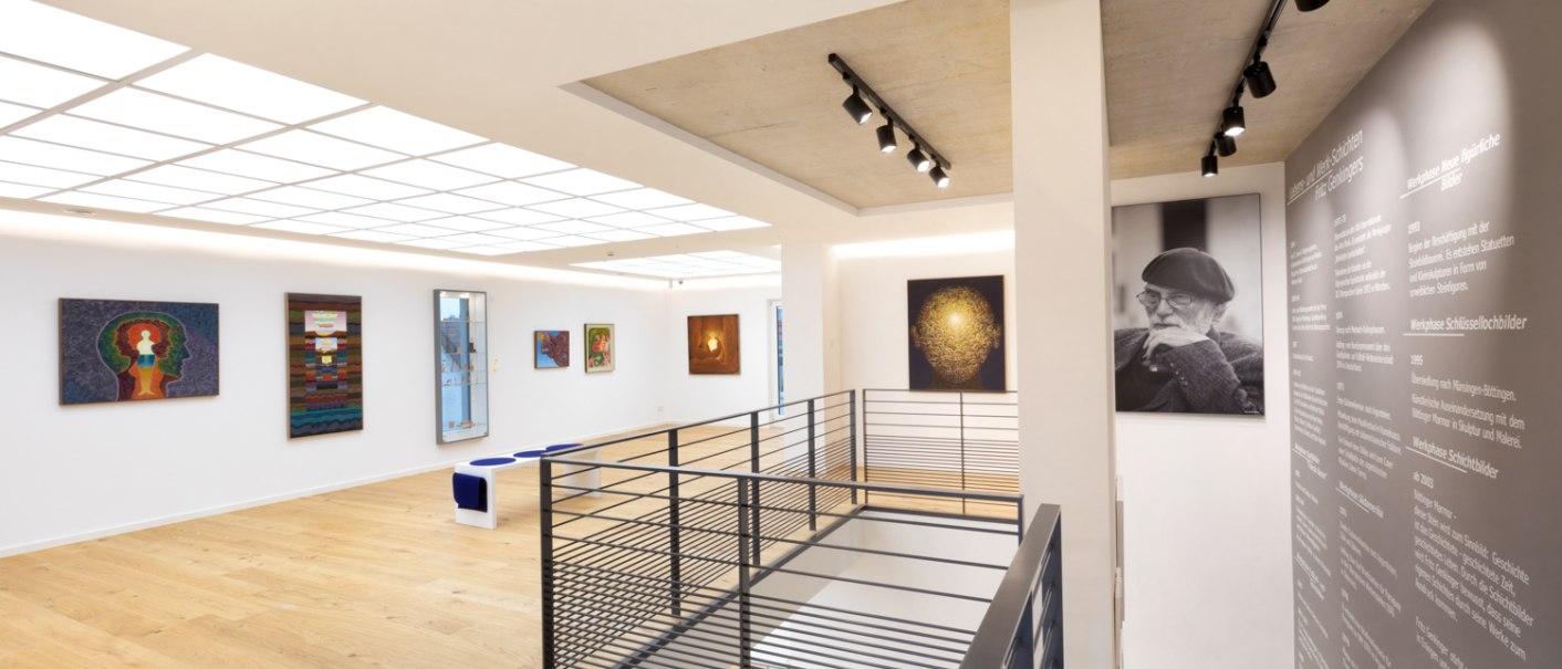 A modern art gallery with paintings on white walls, wooden floors, and a portrait photograph. Lighting from above; a stair railing in the foreground., © Freundeskreis Fritz Genkinger e.V. A modern art gallery with paintings on white walls, wooden floors, and a portrait photograph. Lighting from above; a stair railing in the foreground., © Freundeskreis Fritz Genkinger e.V.