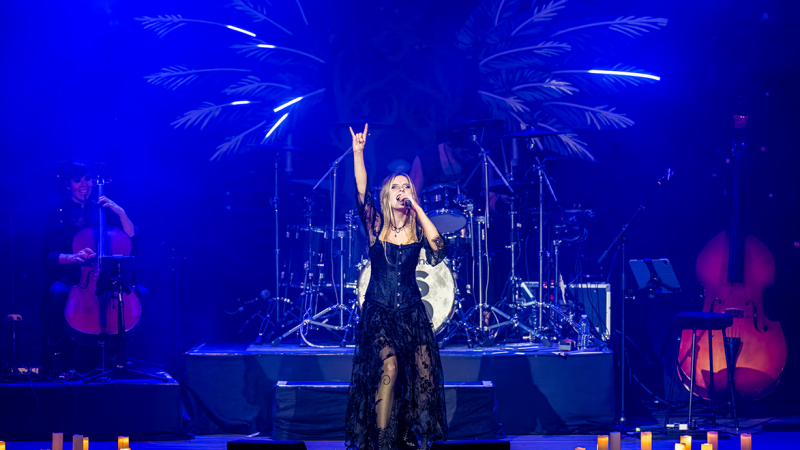 Sängerin in schwarzem Kleid performt auf Bühne, umgeben von Orchesterinstrumenten und Kerzenlicht. Blaue Beleuchtung und dramatische Atmosphäre., © LONDON SYMPHONIC ROCK ORCHESTRA Sängerin in schwarzem Kleid performt auf Bühne, umgeben von Orchesterinstrumenten und Kerzenlicht. Blaue Beleuchtung und dramatische Atmosphäre., © LONDON SYMPHONIC ROCK ORCHESTRA