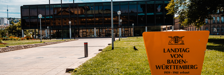 Der Landtag von Baden-Württemberg mit moderner Glasfassade und einem gelben Schild im Vordergrund. Im Hintergrund ist der Fernsehturm zu sehen., © Stuttgart-Marketing GmbH, Sarah Schmid