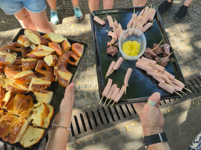 Two trays with pretzels and sausage snacks are presented outdoors. In the background, people are standing on a paved path., © Cool-Tours StattReisen Two trays with pretzels and sausage snacks are presented outdoors. In the background, people are standing on a paved path., © Cool-Tours StattReisen