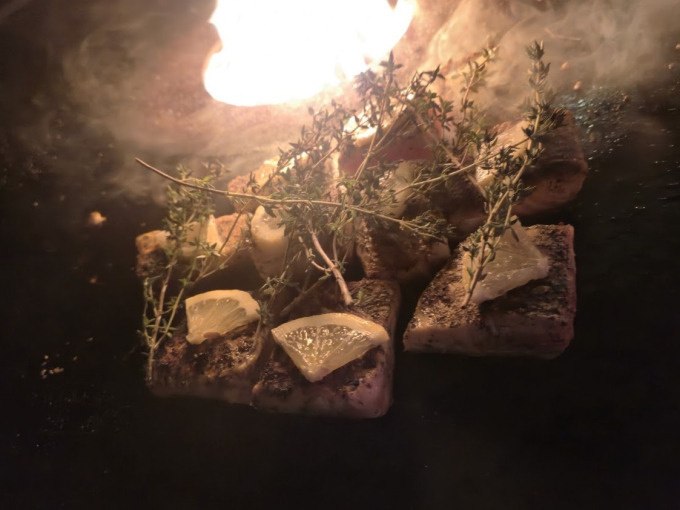 Gegrillter Fisch mit Zitronenscheiben und Kr&auml;utern auf einem Grill. Im Hintergrund lodern Flammen, die eine warme Atmosph&auml;re schaffen., &copy; Cool-Tours StattReisen