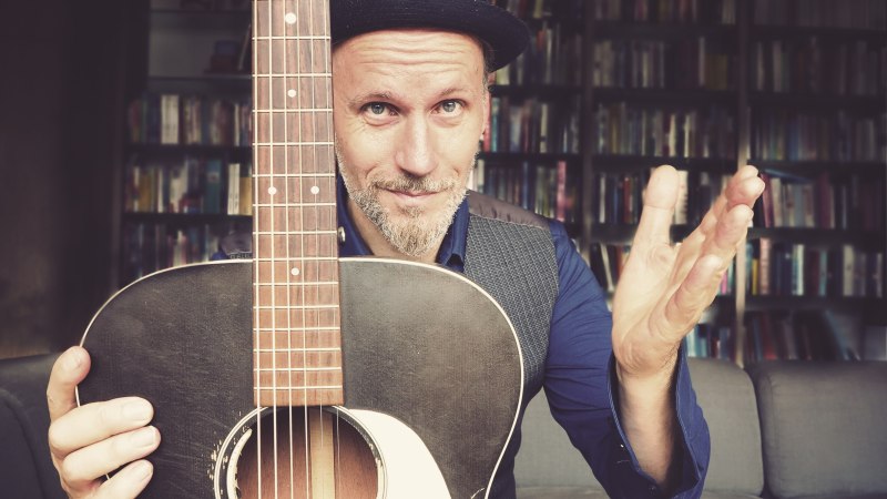 Ein Mann mit Hut hält eine Gitarre vor sich. Im Hintergrund ist ein Bücherregal zu sehen. Er trägt ein blaues Hemd und eine Weste., © Pressefoto Ein Mann mit Hut hält eine Gitarre vor sich. Im Hintergrund ist ein Bücherregal zu sehen. Er trägt ein blaues Hemd und eine Weste., © Pressefoto