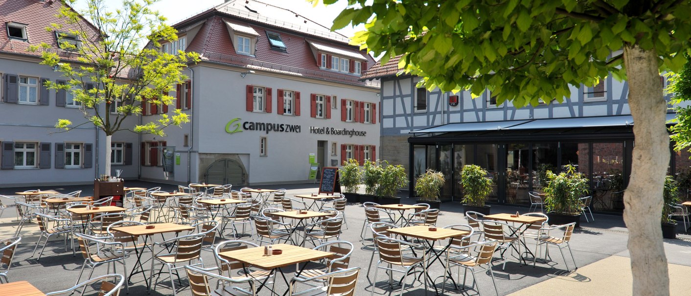Ein sonniger Hotelhof mit leeren Tischen und Stühlen, umgeben von Fachwerkhäusern und grünen Bäumen., © TOMAS