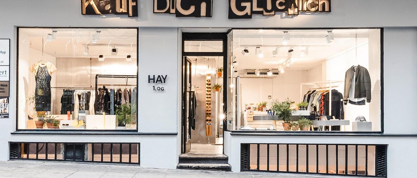 Schaufenster eines Modegeschäfts namens 'Kauf Dich Glücklich' mit Kleidung und Pflanzen dekoriert., © Kauf Dich Glücklich GmbH