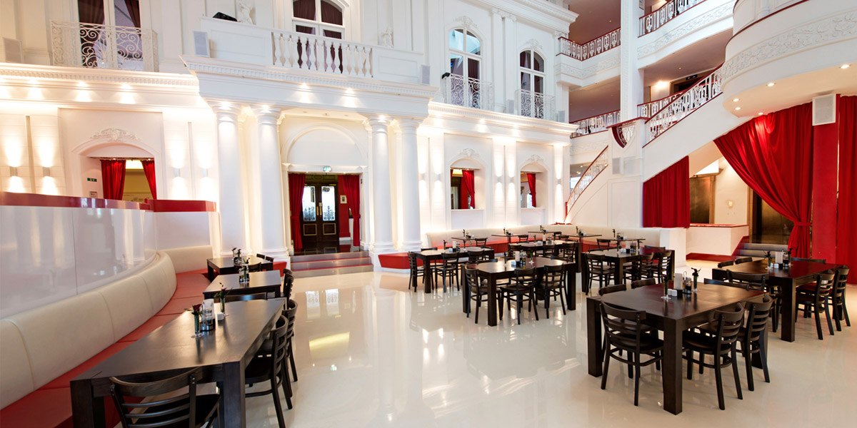 Elegantes Restaurant mit hohen weißen Wänden, roten Vorhängen und dunklen Holztischen. Die Architektur ist klassisch mit Säulen und Balkonen., © Dormero Hotel Stuttgart
