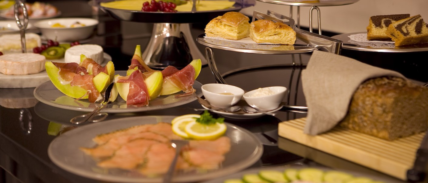 Ein Frühstücksbuffet mit Melone und Schinken, Gebäck, Käse, Lachs, Gemüse und Brot auf einem Tisch arrangiert., © Ross Messehotel Ein Frühstücksbuffet mit Melone und Schinken, Gebäck, Käse, Lachs, Gemüse und Brot auf einem Tisch arrangiert., © Ross Messehotel