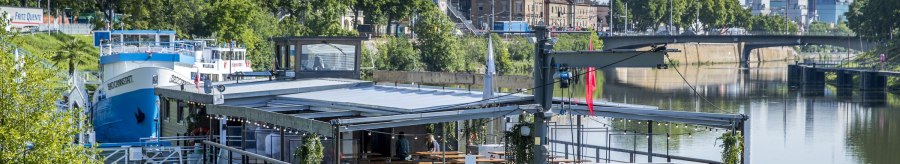A floating restaurant on the Neckar, surrounded by green vegetation and urban buildings in the background., &copy; all copyrights are reserved by maks richter