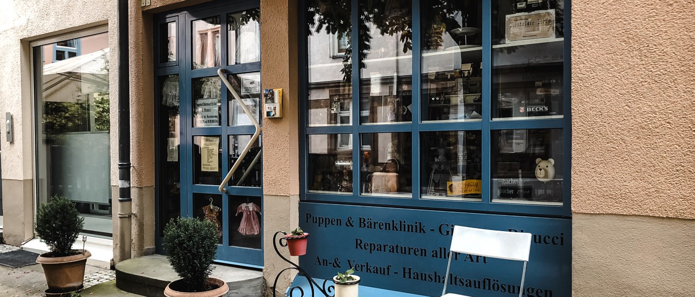 Eingang einer Puppenklinik mit blauer Tür und Schaufenster. Davor stehen Topfpflanzen und ein weißer Stuhl. Ein Schild weist auf Reparaturen hin., © Puppen- und Bärenklinik