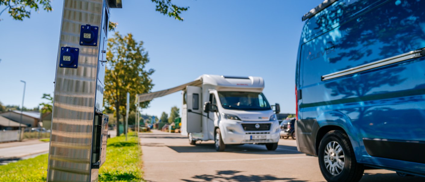 Wohnmobilstellplatz mit einem wei&szlig;en Wohnmobil und einem blauen Van. Im Vordergrund eine Stroms&auml;ule, im Hintergrund B&auml;ume und blauer Himmel., &copy; Stuttgart-Marketing GmbH, Thomas Niederm&uuml;ller