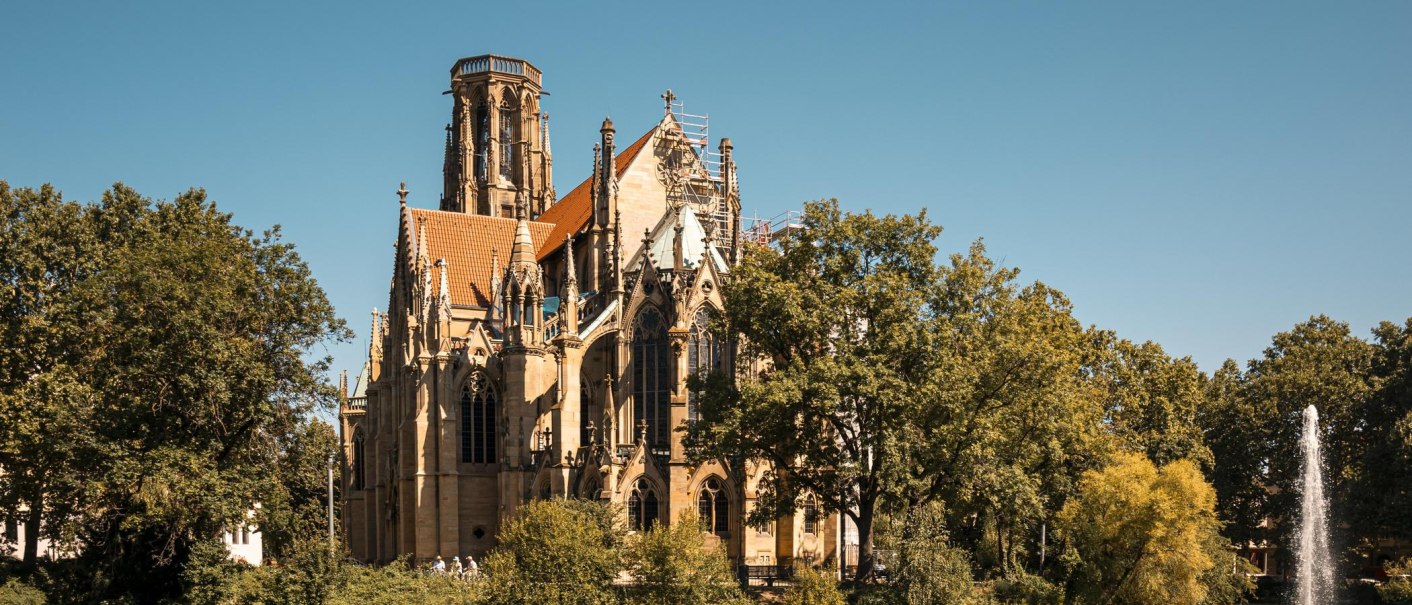 Die Johanneskirche am Feuersee, umgeben von Bäumen, mit einem Springbrunnen im Vordergrund unter klarem Himmel., © SMG, Sarah Schmid