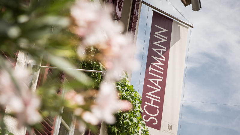 Fahne mit der Aufschrift 'SCHNAITMANN' hängt an einem Gebäude, umgeben von grünen Pflanzen und blühenden Blumen., © Weingut Schnaitmann