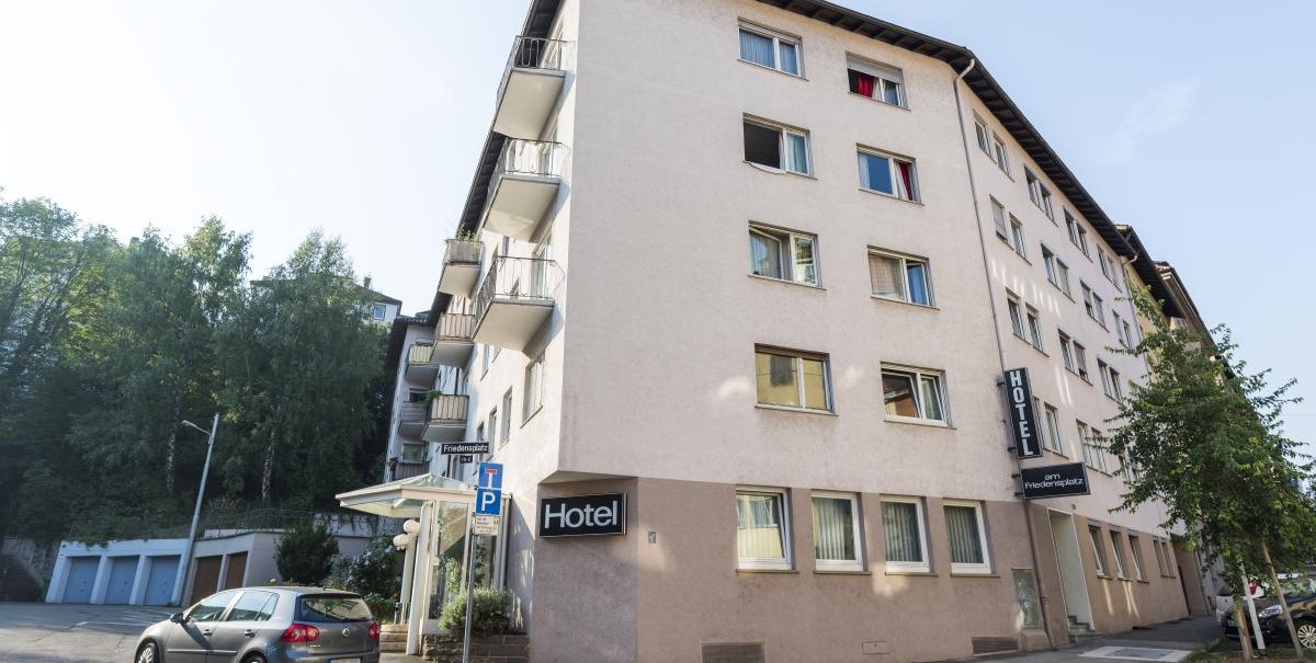 Mehrstöckiges Hotel mit Balkonen, geparktes Auto davor, Straßenschild 'Friedensplatz' sichtbar. Klare, sonnige Wetterbedingungen., © ARNOTEL GmbH