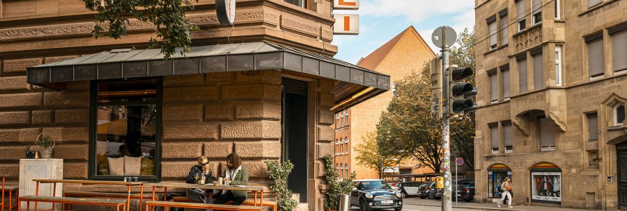 A street corner with a butcher's shop. Two people are sitting at a table in front of the store. A car drives past and there is a traffic light., © SMG, Sarah Schmid