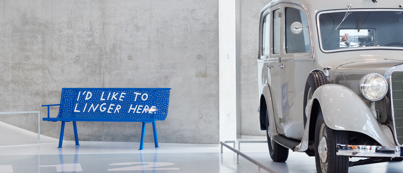 Blue bench with the inscription 'I'd like to linger here' next to a classic car in a modern showroom., © Finnegan Shannon