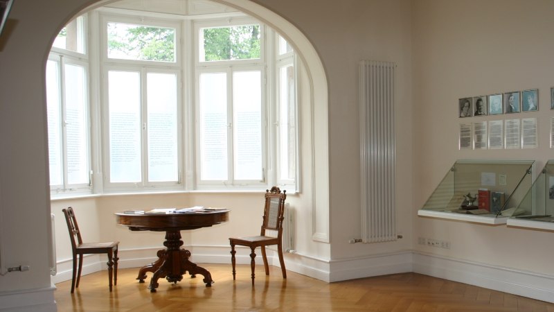 A bright room with a bay window, a round table, and chairs. Photos and texts hang on the wall, and next to them are display cases filled with books., © Gemeindearchiv Bad Boll A bright room with a bay window, a round table, and chairs. Photos and texts hang on the wall, and next to them are display cases filled with books., © Gemeindearchiv Bad Boll