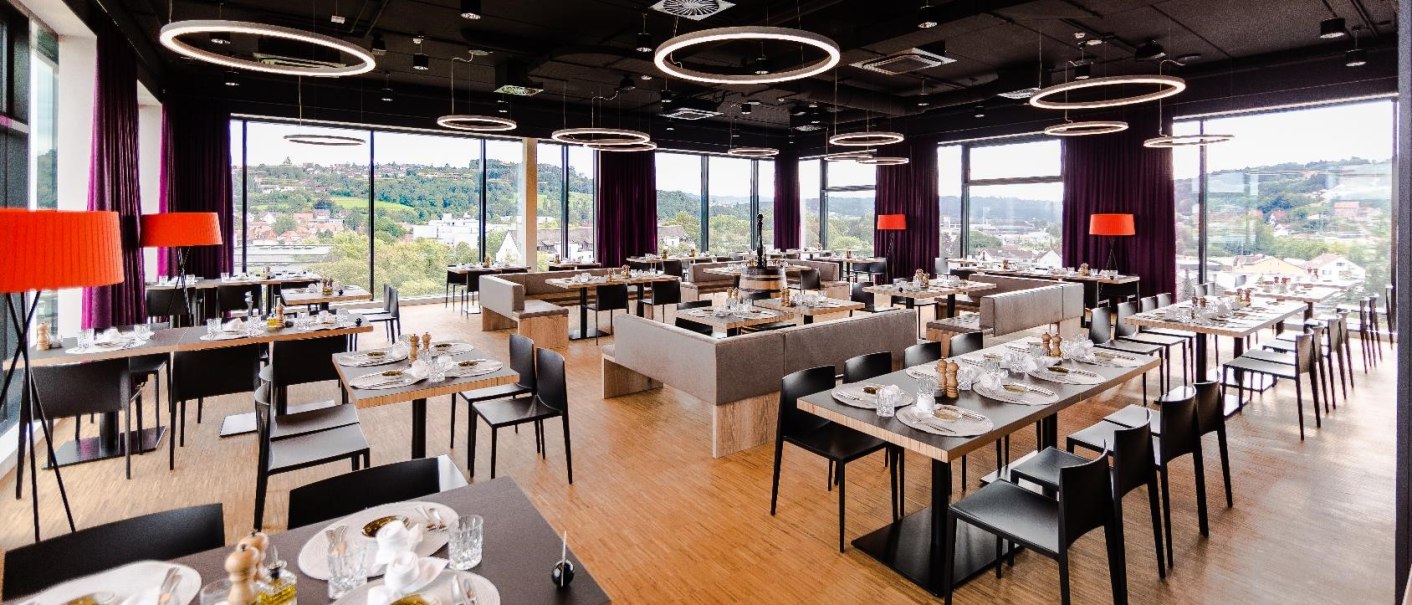 Modern restaurant on the 7th floor with large windows, stylish furnishings and a view of a green landscape. Round ceiling lights and red lamps., © Hotel am Remspark Modern restaurant on the 7th floor with large windows, stylish furnishings and a view of a green landscape. Round ceiling lights and red lamps., © Hotel am Remspark