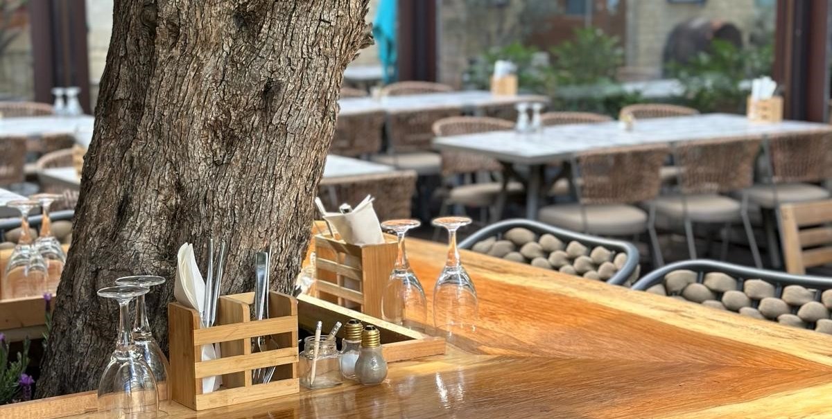 Ein Restauranttisch im Freien mit einem Baum in der Mitte, umgeben von Stühlen. Der Tisch ist gedeckt mit Gläsern und Besteck., © ©Meze Meze