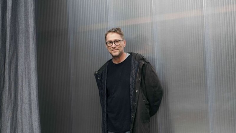 Ein Mann mit Brille und dunklem Mantel lehnt lässig an einer strukturierten Metallwand. Er trägt ein schwarzes Shirt und graue Hose., © Theaterhaus Stuttgart e.V. Ein Mann mit Brille und dunklem Mantel lehnt lässig an einer strukturierten Metallwand. Er trägt ein schwarzes Shirt und graue Hose., © Theaterhaus Stuttgart e.V.