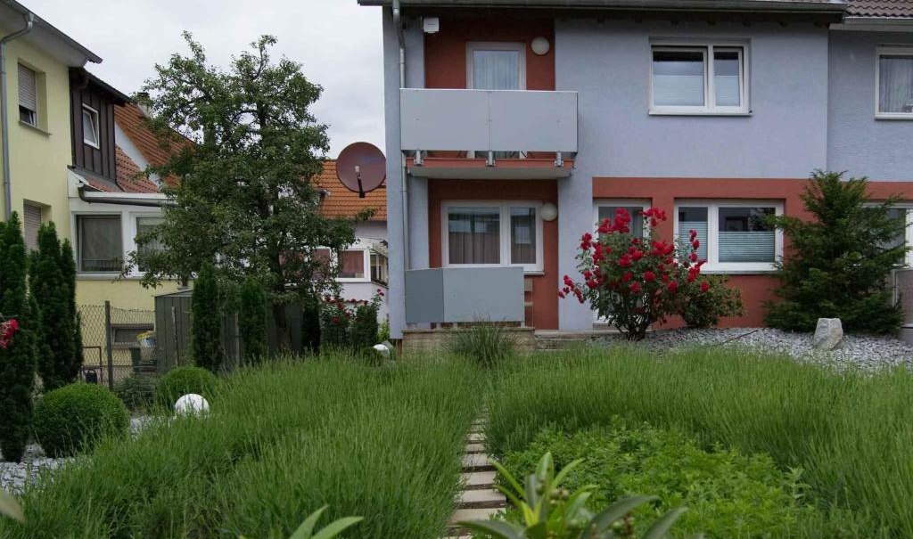 Terraced house with a gray roof, surrounded by a well-kept garden with green bushes and flowering shrubs. A path leads through the garden., © Nödinger Gastro GmbH