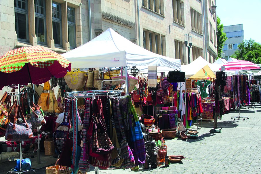 Ein lebhafter Markt mit bunten Ständen, die Kleidung, Taschen und Accessoires anbieten. Weiße und bunte Sonnenschirme spenden Schatten., © Forum der Kulturen Stuttgart e. V. Ein lebhafter Markt mit bunten Ständen, die Kleidung, Taschen und Accessoires anbieten. Weiße und bunte Sonnenschirme spenden Schatten., © Forum der Kulturen Stuttgart e. V.
