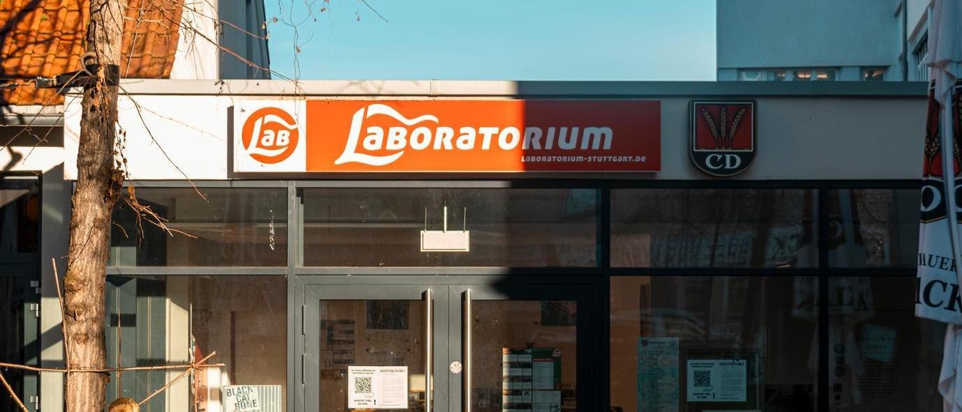 Eingang des Laboratoriums in Stuttgart mit einem markanten roten Schild über der Glasfront. Ein Baum wirft Schatten auf die Fassade., © SMG, Sarah Schmid Eingang des Laboratoriums in Stuttgart mit einem markanten roten Schild über der Glasfront. Ein Baum wirft Schatten auf die Fassade., © SMG, Sarah Schmid