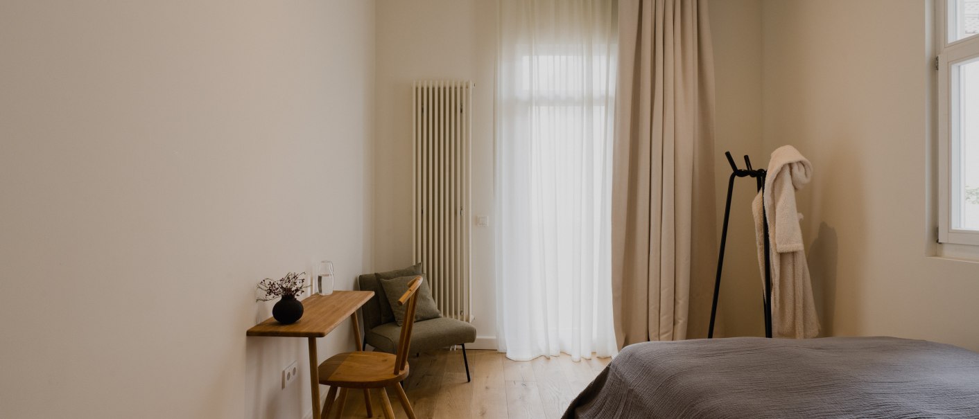 Minimalistisch eingerichtetes Schlafzimmer mit Holzboden, Bett, kleinem Schreibtisch und Stuhl. Vorhänge und ein Bademantel auf einem Ständer ergänzen das Bild., © Peter Oliver Wolff / DAS EDITH