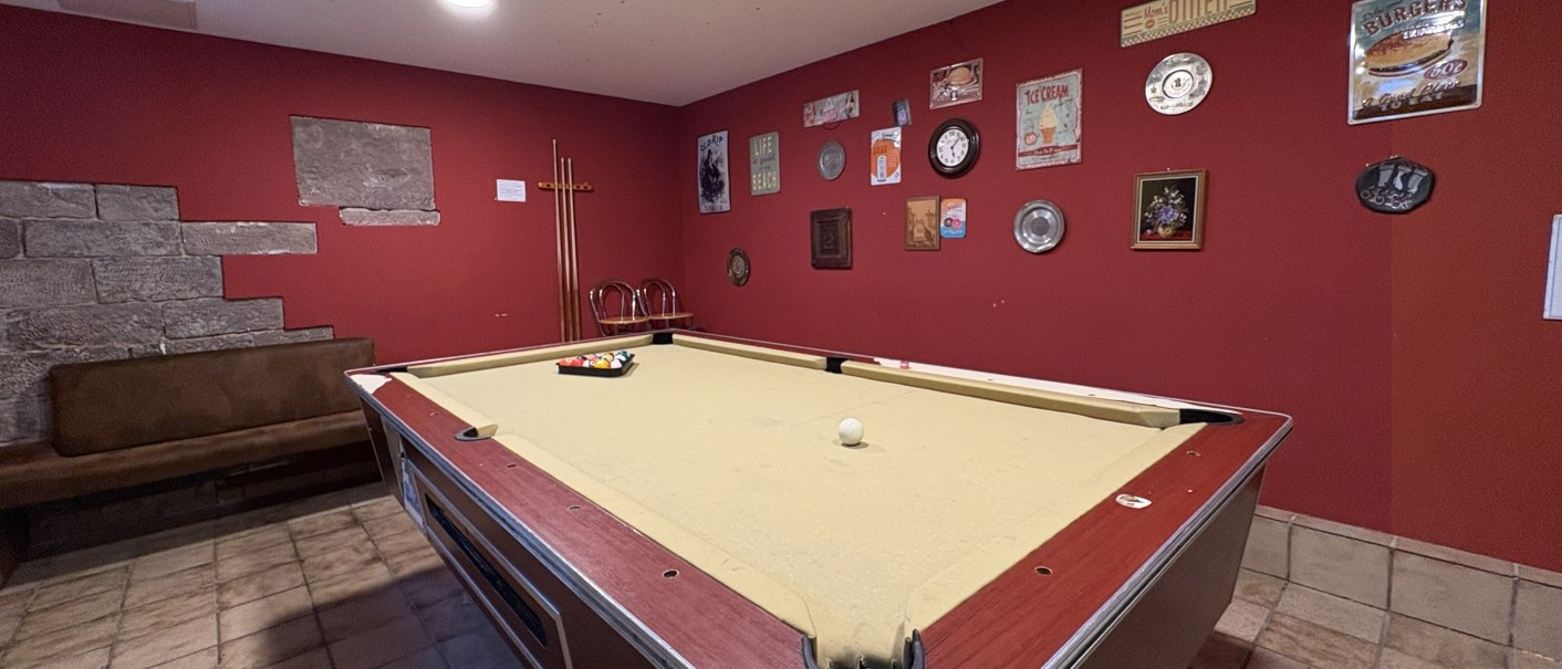 Ein Billardtisch in einem gemütlichen Raum mit roten Wänden, dekoriert mit Vintage-Schildern und einer Uhr. Stühle und ein Queue-Ständer im Hintergrund., © TOMAS Ein Billardtisch in einem gemütlichen Raum mit roten Wänden, dekoriert mit Vintage-Schildern und einer Uhr. Stühle und ein Queue-Ständer im Hintergrund., © TOMAS