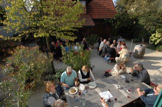 Gem&uuml;tlicher Au&szlig;enbereich mit mehreren Tischen, an denen Menschen essen und trinken. Umgeben von Pflanzen und B&auml;umen, entspannte Atmosph&auml;re., &copy; Elsbeth Koch