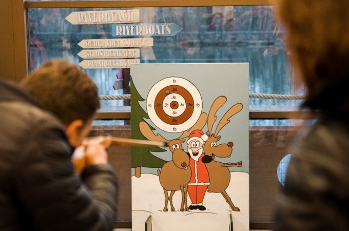 Two people are aiming blowpipes at a target with a Santa Claus and reindeer. Signs and a window can be seen in the background., &copy; Cool-Tours StattReisen