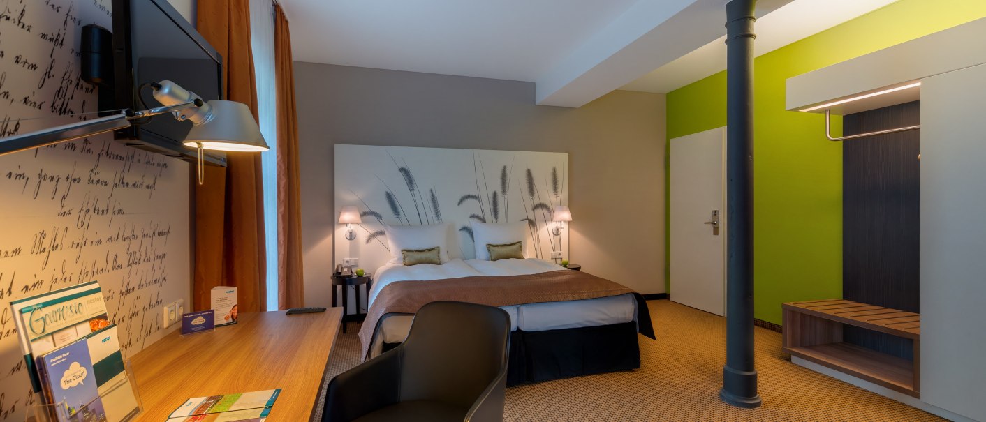 Modernes Hotelzimmer mit Doppelbett, Schreibtisch, Stuhl und Fernseher. Die Wände sind in Grün und Beige gehalten, mit dekorativem Kopfteil., © TOMAS Modernes Hotelzimmer mit Doppelbett, Schreibtisch, Stuhl und Fernseher. Die Wände sind in Grün und Beige gehalten, mit dekorativem Kopfteil., © TOMAS