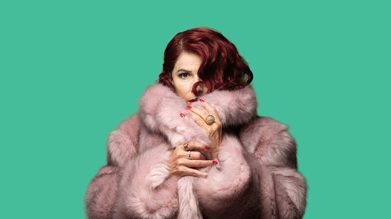 Eine Frau mit rotem Haar trägt einen rosa Pelzmantel und hält ihn nah am Gesicht. Der Hintergrund ist einfarbig grün., © Württembergische Staatstheater Stuttgart Eine Frau mit rotem Haar trägt einen rosa Pelzmantel und hält ihn nah am Gesicht. Der Hintergrund ist einfarbig grün., © Württembergische Staatstheater Stuttgart