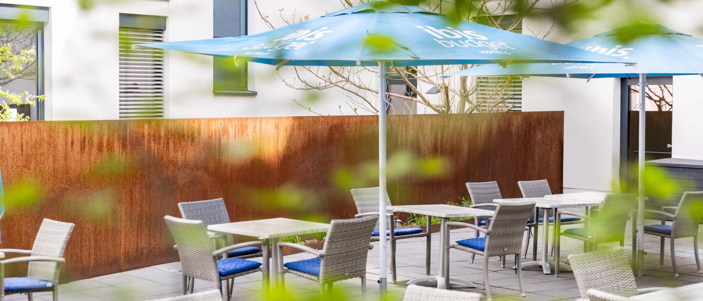 Moderne Terrasse mit geflochtenen Stühlen, Tischen und blauen Sonnenschirmen. Im Hintergrund ein Gebäude mit Fenstern und einer rostfarbenen Wand., © DQuadrat Living GmbH Moderne Terrasse mit geflochtenen Stühlen, Tischen und blauen Sonnenschirmen. Im Hintergrund ein Gebäude mit Fenstern und einer rostfarbenen Wand., © DQuadrat Living GmbH