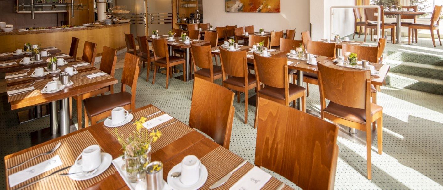 A bright breakfast room with wooden tables and chairs, set with crockery and flowers. A buffet area in the background., © Hotel Gloria GmbH Restaurant Möhringer Hexle A bright breakfast room with wooden tables and chairs, set with crockery and flowers. A buffet area in the background., © Hotel Gloria GmbH Restaurant Möhringer Hexle