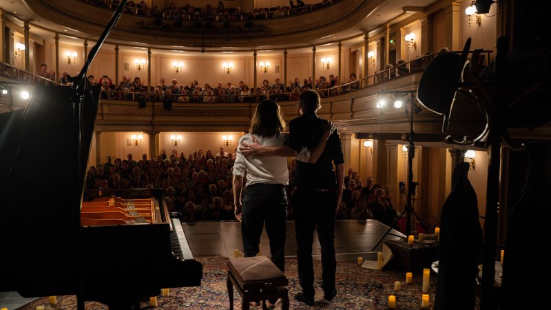 Zwei Künstler verbeugen sich auf einer Bühne vor einem vollen Theater. Ein Flügel steht im Vordergrund, Kerzen und ein Teppich schmücken die Bühne., © Künstler Zwei Künstler verbeugen sich auf einer Bühne vor einem vollen Theater. Ein Flügel steht im Vordergrund, Kerzen und ein Teppich schmücken die Bühne., © Künstler