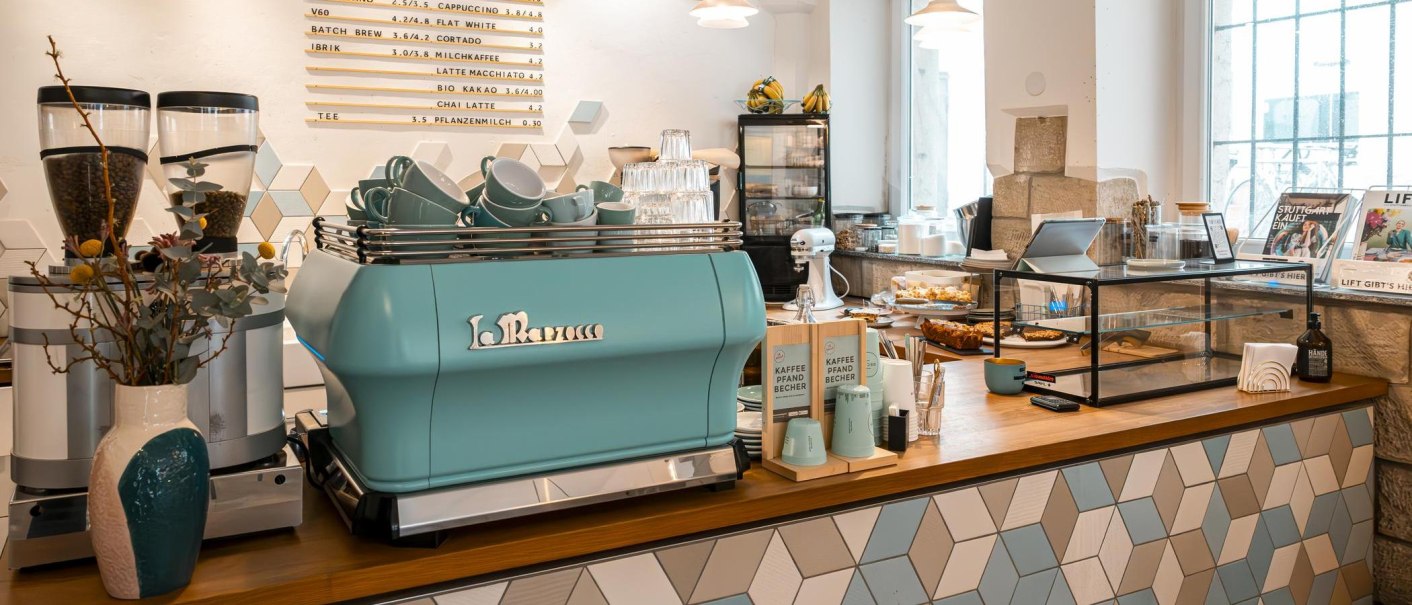 Modernes Café mit blauer Kaffeemaschine, dekorativen Fliesen und Backwaren. Helle, einladende Atmosphäre mit Pflanzen und Zeitschriften., © Stuttgart-Marketing GmbH, Sarah Schmid