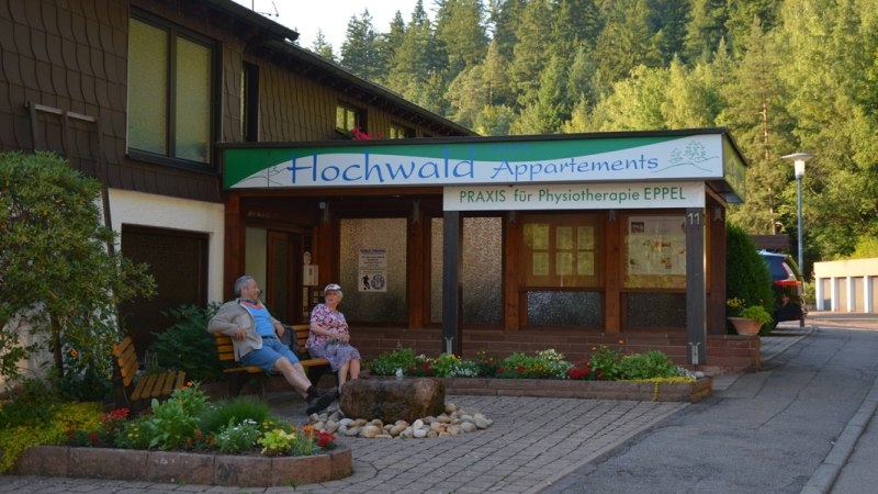 Building with sign 'Hochwald Appartements' and 'Praxis für Physiotherapie EPPEL'. Two people are sitting on a bench, surrounded by plants and trees., © Hotel Hochwald Building with sign 'Hochwald Appartements' and 'Praxis für Physiotherapie EPPEL'. Two people are sitting on a bench, surrounded by plants and trees., © Hotel Hochwald