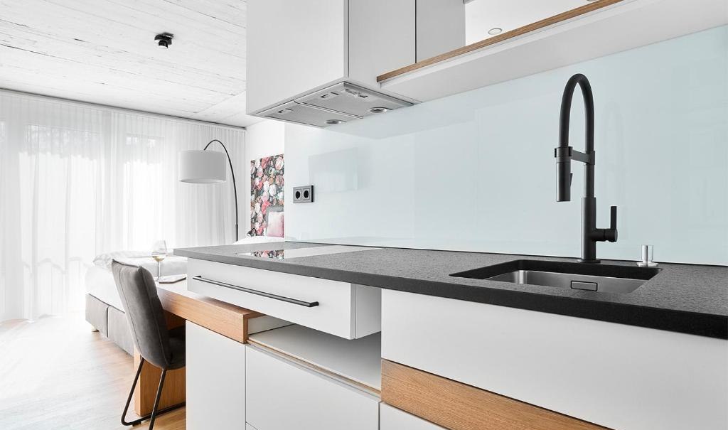 Moderne Kochnische mit weißem Schrank, schwarzer Arbeitsplatte und Wasserhahn. Im Hintergrund ein heller Wohnbereich mit Sofa und Stehlampe., © livisit bergapartments