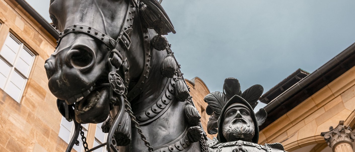 Nahaufnahme einer Reiterstatue im Innenhof eines alten Schlosses. Der Reiter trägt eine Rüstung und einen Federhelm, das Pferd ist detailliert verziert., © Stuttgart Marketing GmbH - Sarah Schmid