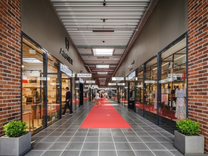 Eine moderne Einkaufspassage mit roten Teppichen und Geschäften wie Aigner auf beiden Seiten. Menschen schlendern durch den Gang., © Schlienz-Tours GmbH & Co.KG Eine moderne Einkaufspassage mit roten Teppichen und Geschäften wie Aigner auf beiden Seiten. Menschen schlendern durch den Gang., © Schlienz-Tours GmbH & Co.KG