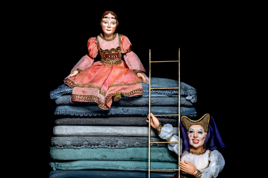 Zwei Puppen in historischen Kostümen: Eine sitzt auf einem hohen Stapel Matratzen, die andere hält eine Leiter. Der Hintergrund ist schwarz., © Schäfer & Thieme Produktion Zwei Puppen in historischen Kostümen: Eine sitzt auf einem hohen Stapel Matratzen, die andere hält eine Leiter. Der Hintergrund ist schwarz., © Schäfer & Thieme Produktion