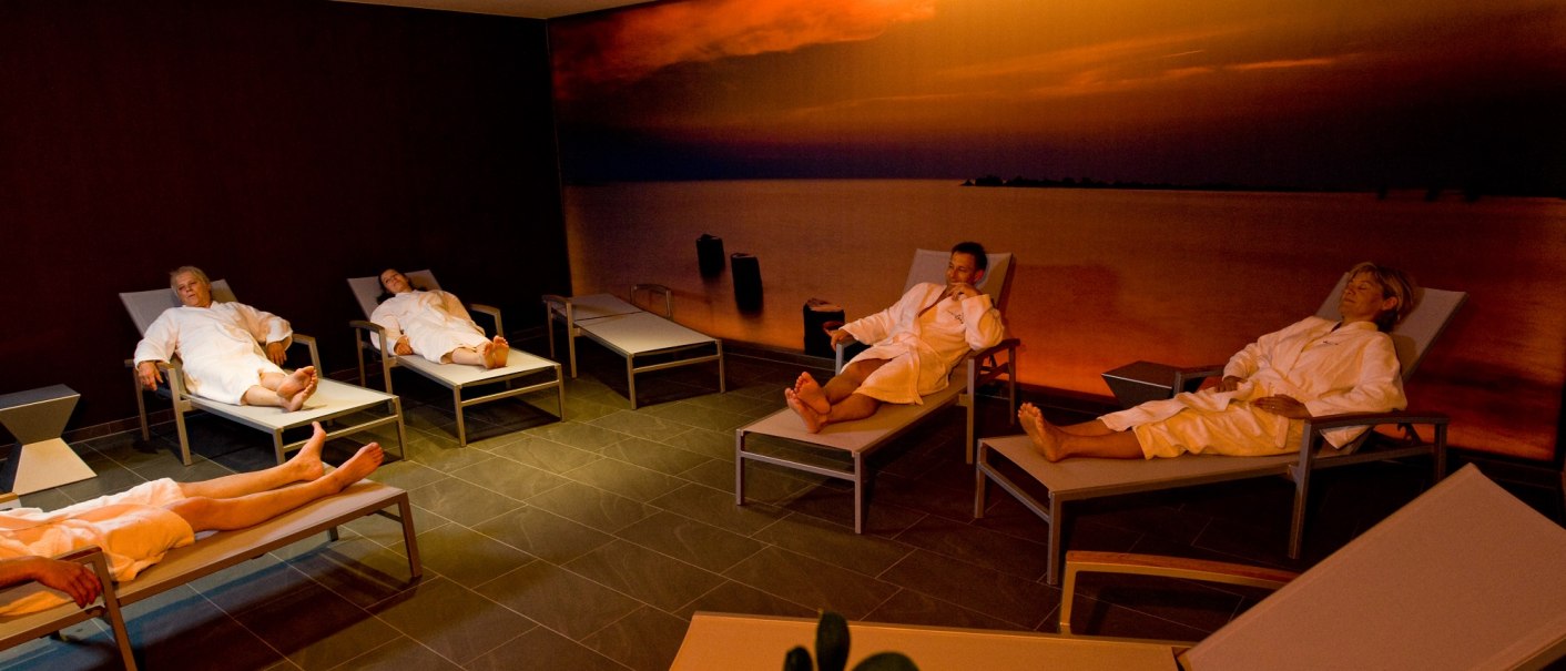 Personen in Bademänteln entspannen auf Liegen in einem Spa-Raum. Die Wand zeigt ein Bild eines Sonnenuntergangs über dem Meer., © Hotel Fortuna