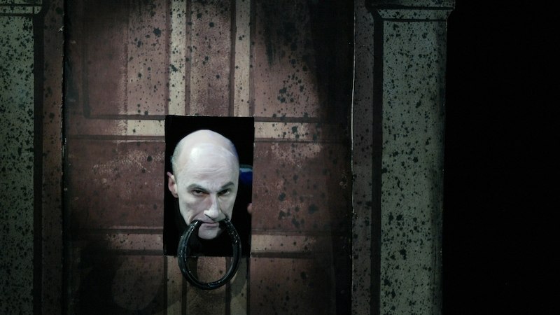 Ein Mann blickt durch ein kleines Fenster in einer alten Holztür mit einem großen, runden Türklopfer., © Wilhelma Theater Ein Mann blickt durch ein kleines Fenster in einer alten Holztür mit einem großen, runden Türklopfer., © Wilhelma Theater