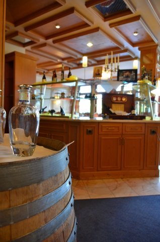 Innenansicht eines Restaurants mit Holzmöbeln, einer Theke und dekorativen Flaschen. Ein Weinfass dient als Tisch mit einer Karaffe darauf., © Badhotel Restaurant Stauferland