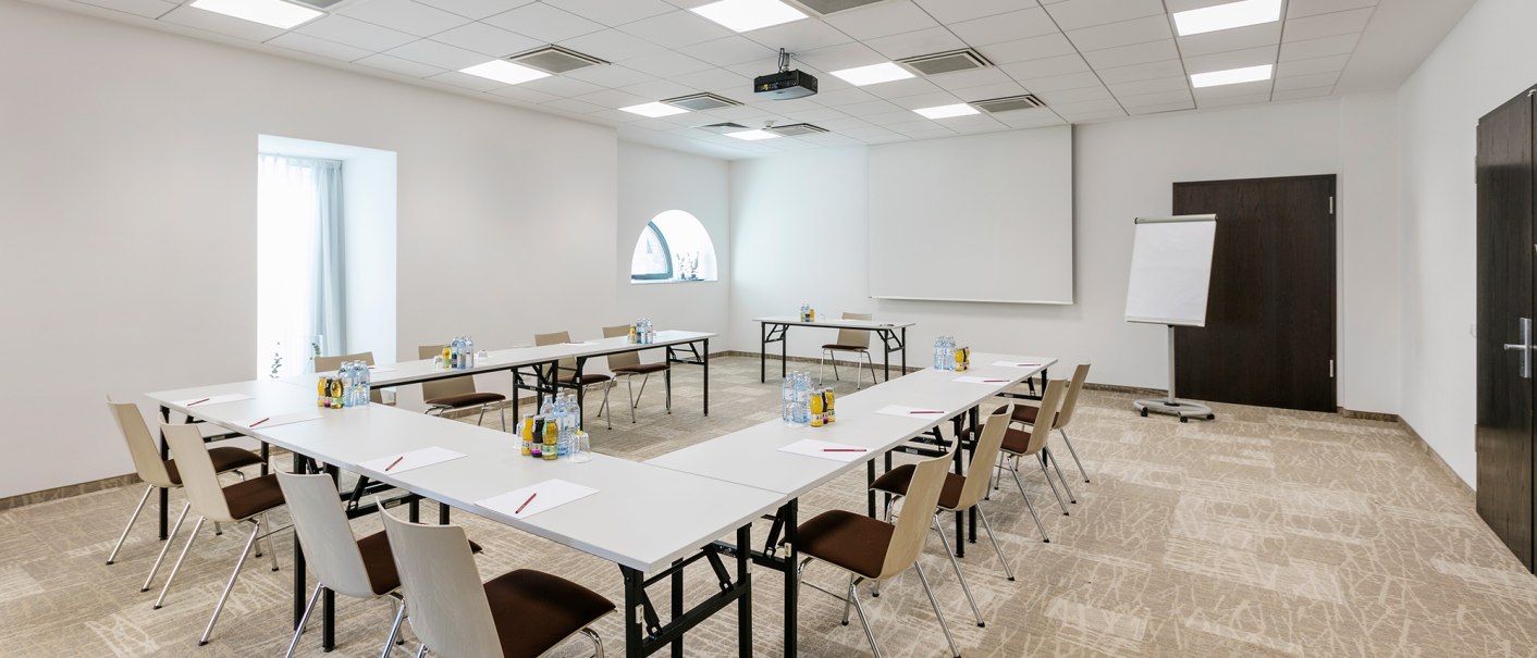 Modern conference room with U-shaped table arrangement, chairs, flipchart and projector. Bright lighting and neutral colors dominate the room., © Arcotel Hotels Modern conference room with U-shaped table arrangement, chairs, flipchart and projector. Bright lighting and neutral colors dominate the room., © Arcotel Hotels