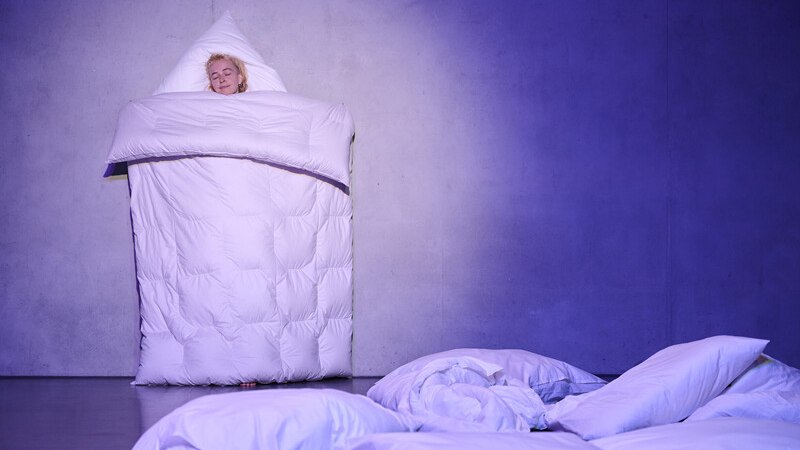 Eine Person steht in eine Decke eingewickelt an einer Wand, umgeben von Kissen auf dem Boden. Die Szene ist in blauem Licht getaucht., © Württembergische Staatstheater Stuttgart Eine Person steht in eine Decke eingewickelt an einer Wand, umgeben von Kissen auf dem Boden. Die Szene ist in blauem Licht getaucht., © Württembergische Staatstheater Stuttgart